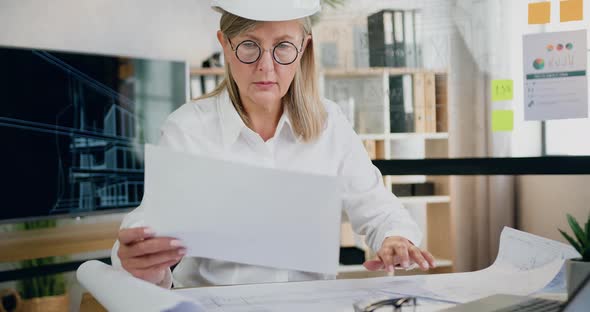 Female Engineer in Helmet Checks Reports and Ready Blueprint of Future Building alt