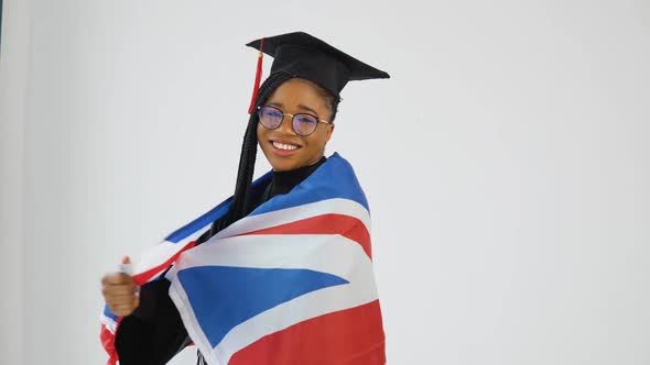 Happy Stylish Afro American Woman Student in Graduate Uniform is Posing to the Camera Holding UK alt