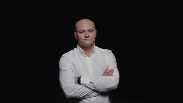 A bald man in a white shirt crosses his arms and poses for the camera on a black background. alt