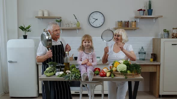 Senior Woman and Man with Grandchild Girl Making a Funny Dance with Strainer and Vegetables at Home alt