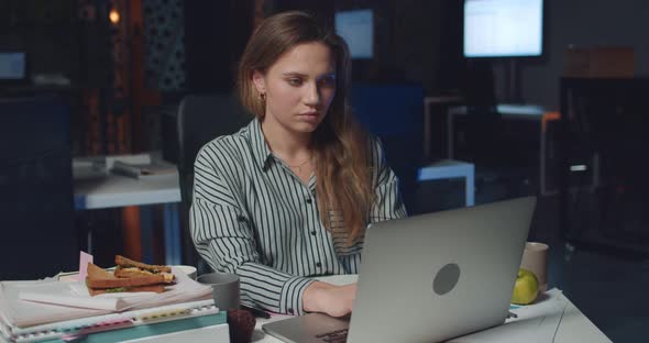 Front View of Pretty Female Office Worker Typing on Her Laptop Late at Night. Young Woman Using Her alt