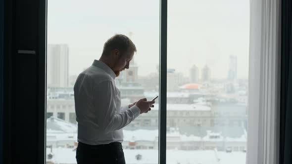 businessman uses smartphone while standing by the window with city view