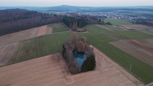 A small lake within green and brown fields in Hesse, Germany. Aerial approach with gimbal tilting do alt