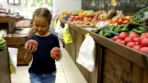 Kid Girl with Two Peaches Is Walking Along Grocery Rows Toward To Cashier