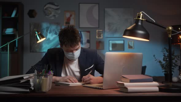 Young Asian Man Wearing Medical Mask Finishing Working Remotely at Home During Coronavirus Lockdown alt