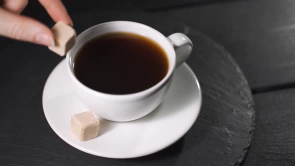 Close Up of a Woman Hand Throwing Sugar Cube Into a Coffee Mug on a Table at Home alt