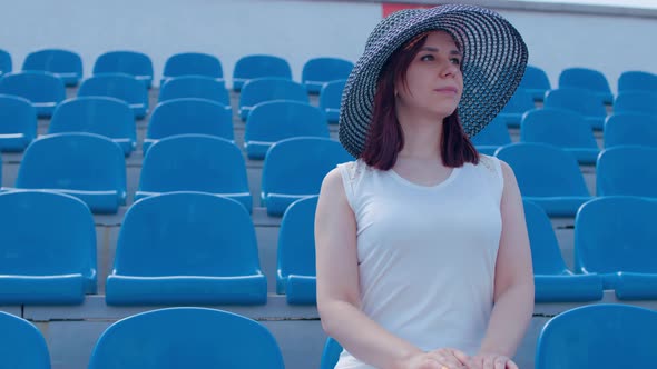 Young Woman in in White Dress and Elegant Hat Looks Away Sitting on Stadium Bleachers Alone alt