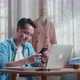 Asian Male Designer With Sewing Machine Looking At Smartphone While Designing Clothes On Laptop - VideoHive Item for Sale