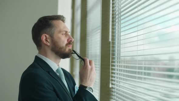 Focused Businessman in a Suit Stands at the Window in the Office and Reflects the Thought Process alt
