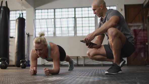 Fit caucasian woman performing plank exercise with male trainer holding digital tablet at the gym alt