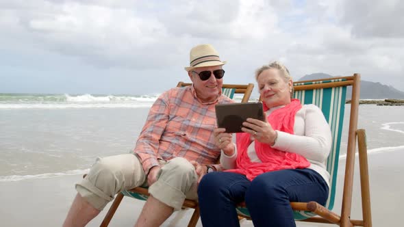Front view of old caucasian senior couple using digital tablet and sitting on sunlounger a beach 4k alt
