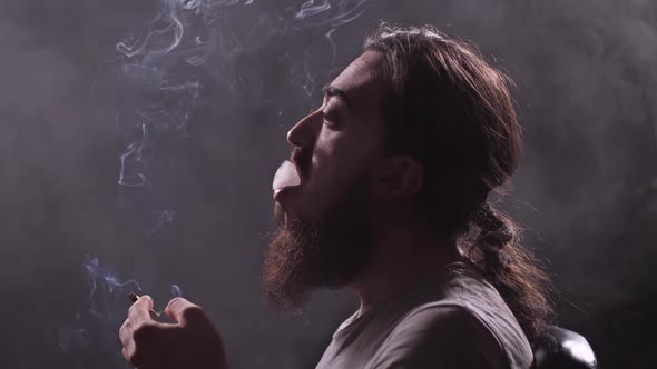 A Man with a Beard and Long Hair Smokes a Shoal in a Dark Room a Lot of Smoke Around Him alt