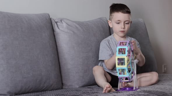 A boy in a gray T-shirt sits on a bed near a white wall and plays with a Ferris wheel alt