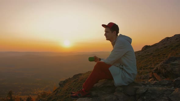 Hipster Man Sitting on Edge of Rock Cliff Drinks Tea From Mug Enjoying Sunset
