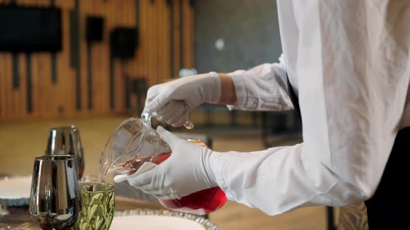 Close Up Waiter Hands in White Gloves Pouring Red Drink From Jug Into Glass Goblet in Restaurant alt