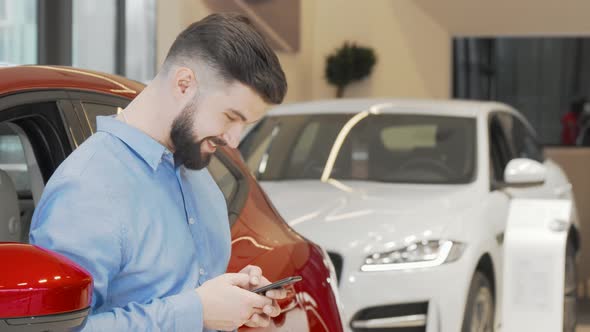 Happy Man Texting on a Smart Phone While Shopping for New Car at the Dealership