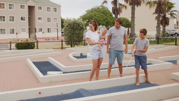 Family of Four Playing on Miniature Golf Course Boy Making Putt Into Hole alt