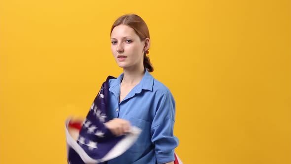 Young Beautiful Woman in Bright Clothes on a Pastel Yellow Background Holds and Develops a USA Flag alt