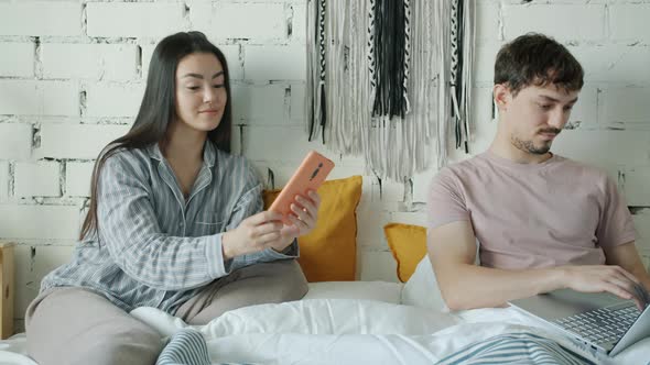 Girl and Guy Using Smartphone and Laptop Relaxing in Bed at Home alt