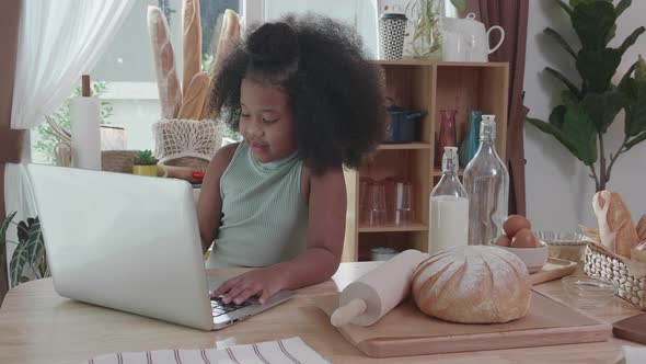 Little adorable afro African American girl learning online by laptop, at table in kitchen