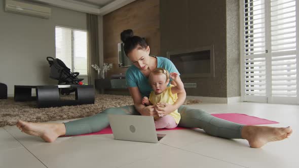 Caucasian mother holding her baby practicing yoga on yoga mat in front of laptop at home alt