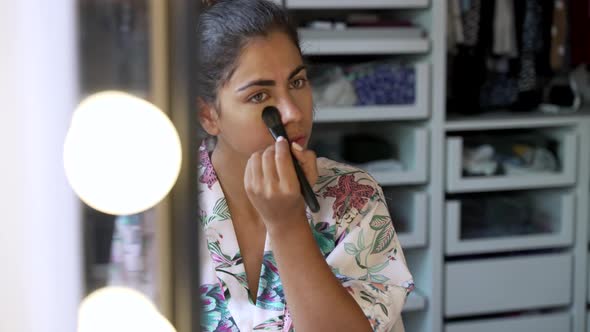 Young woman applying makeup under eyes in bedroom alt