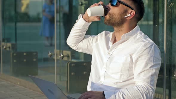 Bearded Middleaged Man in Sunglasses Sits on the Street with a Laptop and Drinks Coffee From a