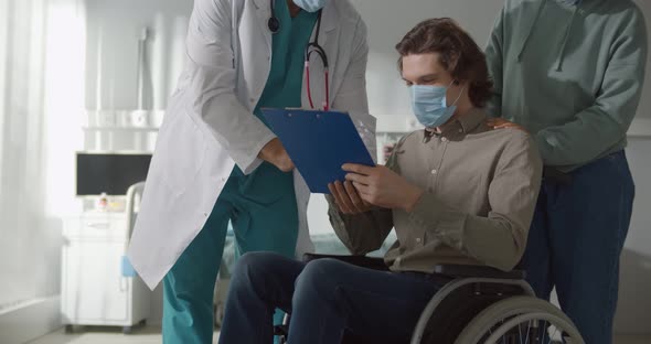 Male Patient Sitting in Wheelchair and Wearing Safety Mask Signing Discharge Paper in Hospital alt