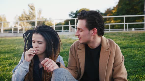 Happy Young Hipster Couple Sitting on Grass and Smoking Marijuana Joint in Park Slow Motion alt