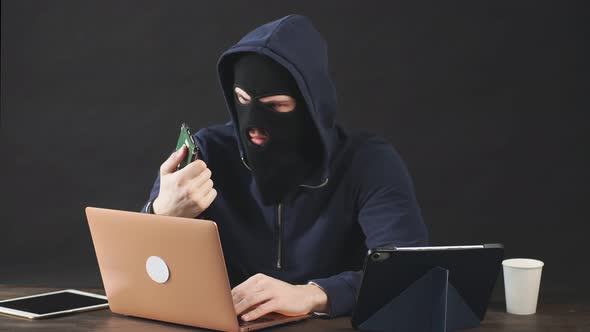 Male Hacker in Black Mask and Hood Uses Laptop To Hack a Hard Drive. alt