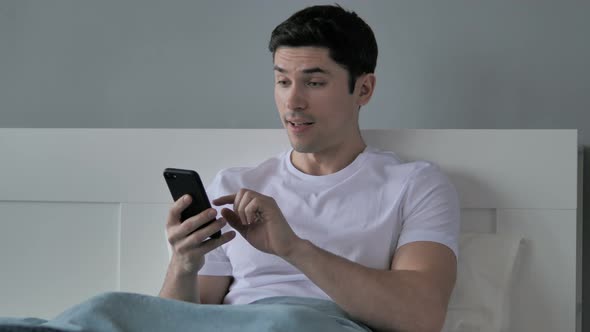 Young Man Reacting To Success, Using Smartphone in Bed alt
