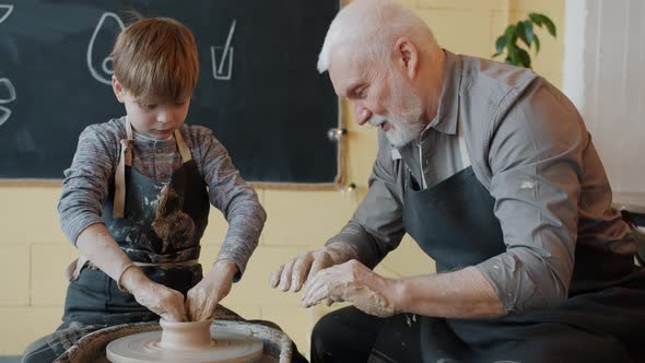 Experienced Potter Teaching Boy To Work with Spinning Wheel in Workshop Speaking alt