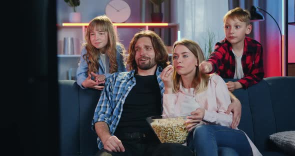 Smiling Parents and Their Two Kids Watching TV-Show at Home in the Evening and Eating Popcorn alt