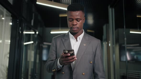 A Black Man in a Business Suit Walks Down the Corridor of the Office Looking at His Mobile Phone alt