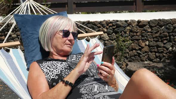 Senior Woman Looking on Cell Phone Relaxing on a Hammock in Home Garden