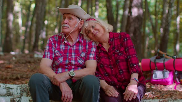 Grandmother Grandfather Senior Tourists Hikers Sitting on Tree and Hugging Kissing in Summer Wood alt