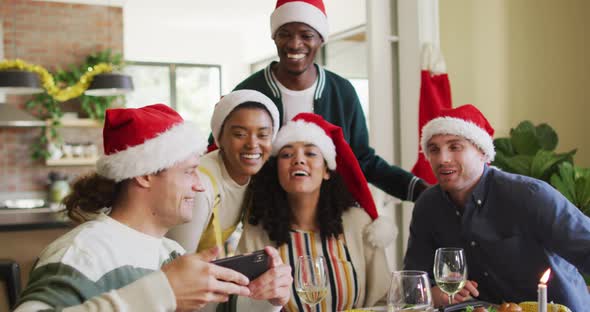 Happy group of diverse friends in santa hats celebrating meal, taking selfie at christmas time alt