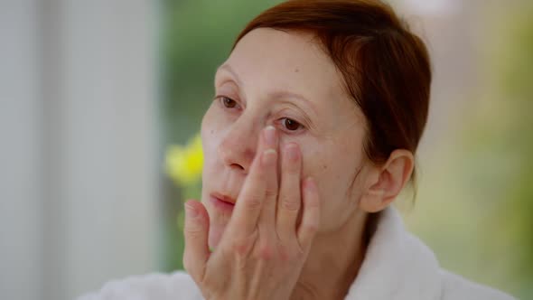 Headshot of Confident Mature Woman Applying Moisturizing Cream on Face Looking Away alt