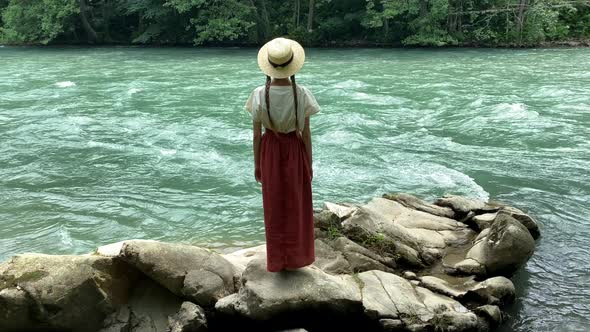 Young vintage woman in dress and straw boater hat stand near rippling mountain river, back view alt