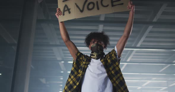 Mixed race man wearing face mask holding protest placard in empty parking garage alt