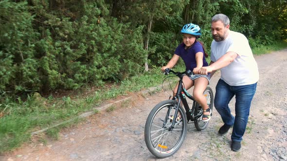 Bearded dad teaches his daughter in a Bicycle helmet to ride a bike in the Park. 