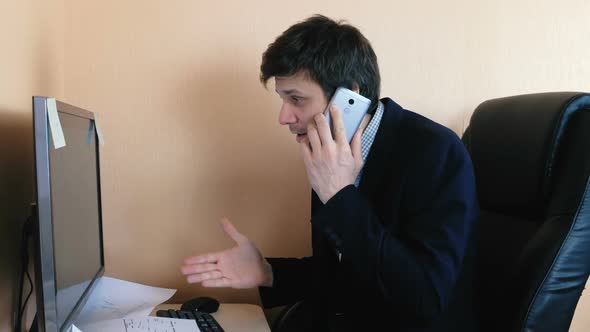 Man Working at the Computer and Calling Mobile Phone. Disgruntled Young Businessman.