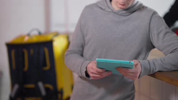 Unrecognizable Male Student Surfing Social Media on Tablet with Yellow Food Delivery Backpack at alt
