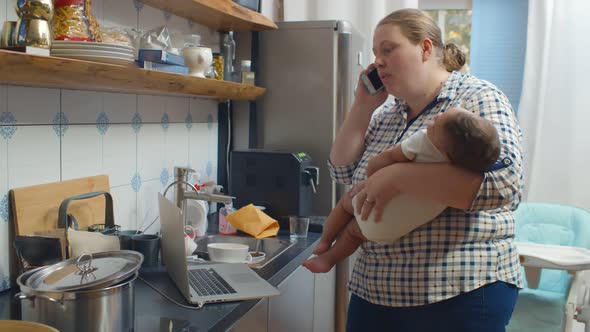 Young Mother Talking on Phone Holding Sleeping Baby at Kitchen alt