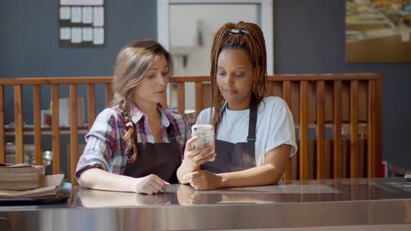 Young Multiethnic Owners of Small Modern Coffee House Using Smartphone for Work. alt