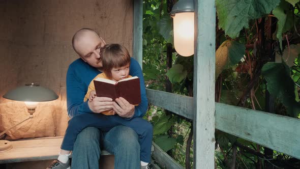 Man Reads a Paper Book for Small Child Sitting on the Porch of a Village House alt