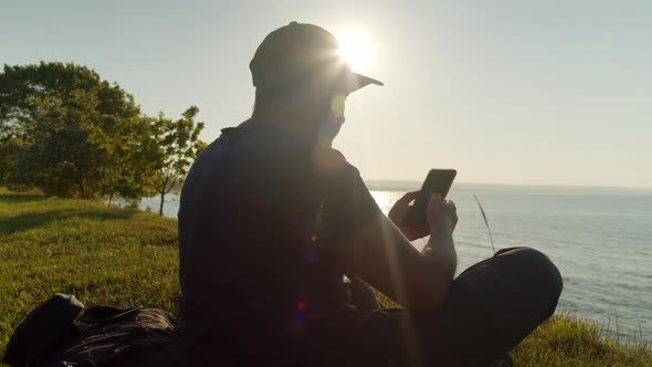 Man Tourist is Resting on the Seashore with a Smartphone in His Hand alt