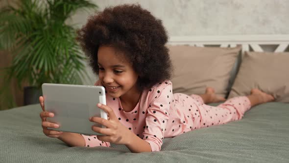 Cute Little African American Girl in Plays a Game on a Digital Tablet and Rejoices at the Victory alt