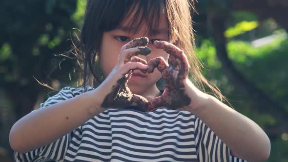Cute little girl with muddy hands makes a heart shape with her hands against nature background. alt