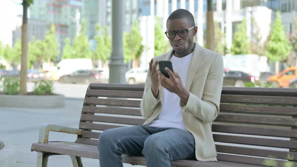 African Man Reacting to Loss on Smartphone While Sitting Outdoor on Bench alt
